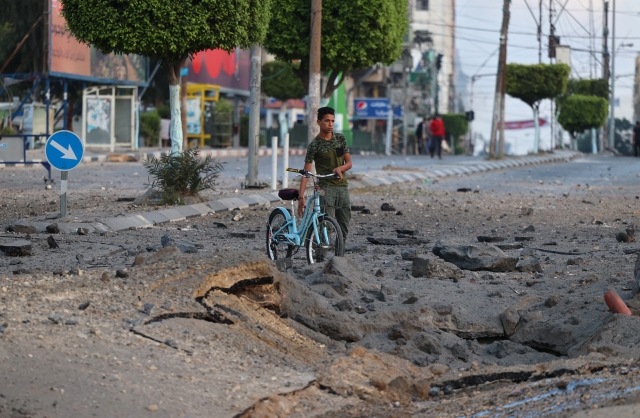 A Palestinian man pushes his bicycle past a crater on the ground near the al-Sharouk tower, which housed the bureau of the Al-Aqsa television channel in the Hamas-controlled Gaza Strip, after it was destroyed by an Israeli air strike, in Gaza City, on May 13, 2021. © Mohammed Abed / AFP